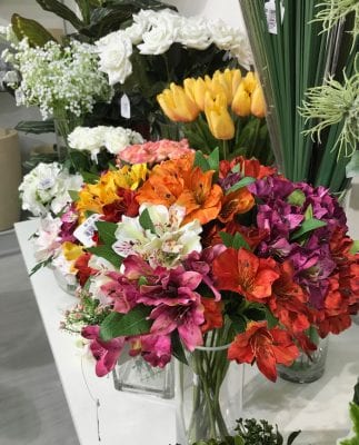 conjunto de flor de azalea artificial em vaso de vidro transparente. No fundo um conjunto de mosquitinho, rosas brancas, tulipas amarela e capim artificial sobre mesa branca.